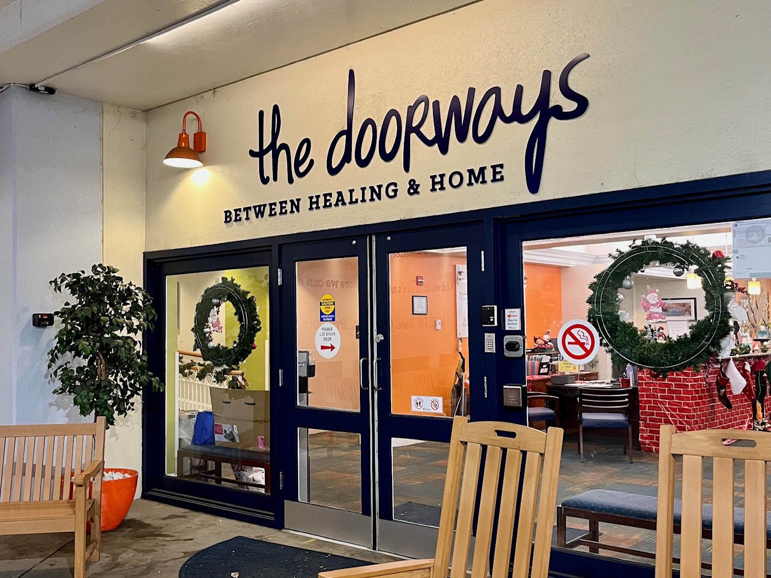 Entrance of The Doorways, flanked by benches and rocking chairs, with holiday wreaths hung in the background. Signage over the door reads "The Doorways, Between Healing & Home."